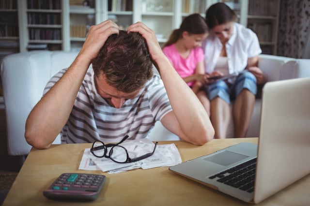 Worried man sitting at table with bills and laptop while his wife and daughter sitting on sofa.jpeg